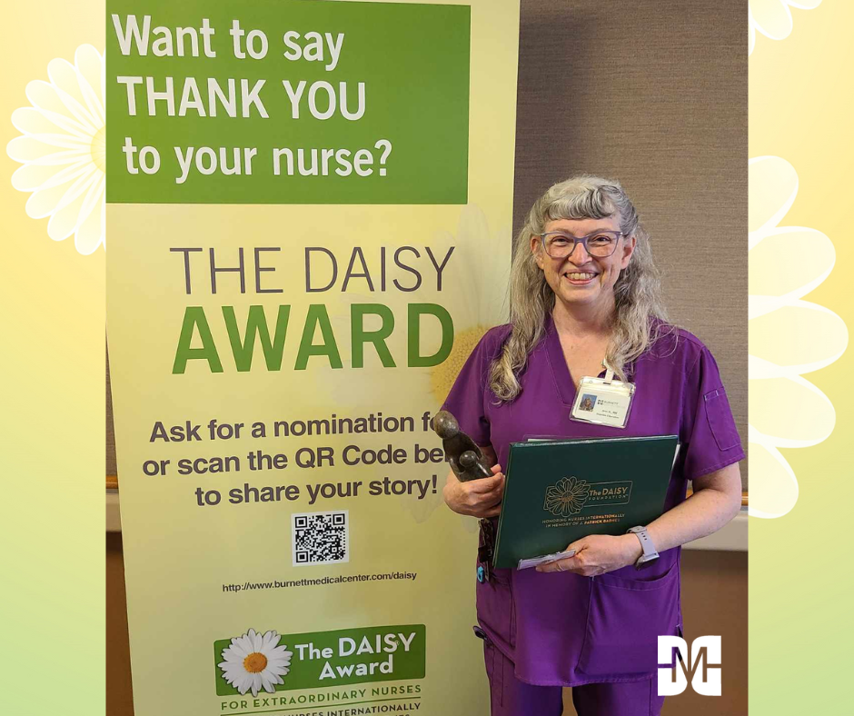 Nurse in purple scrubs holding a DAISY Award nomination form and a decorative hammer, standing next to a banner promoting the DAISY Award for extraordinary nurses at Burnett Medical Center, encouraging appreciation for compassionate nursing care.