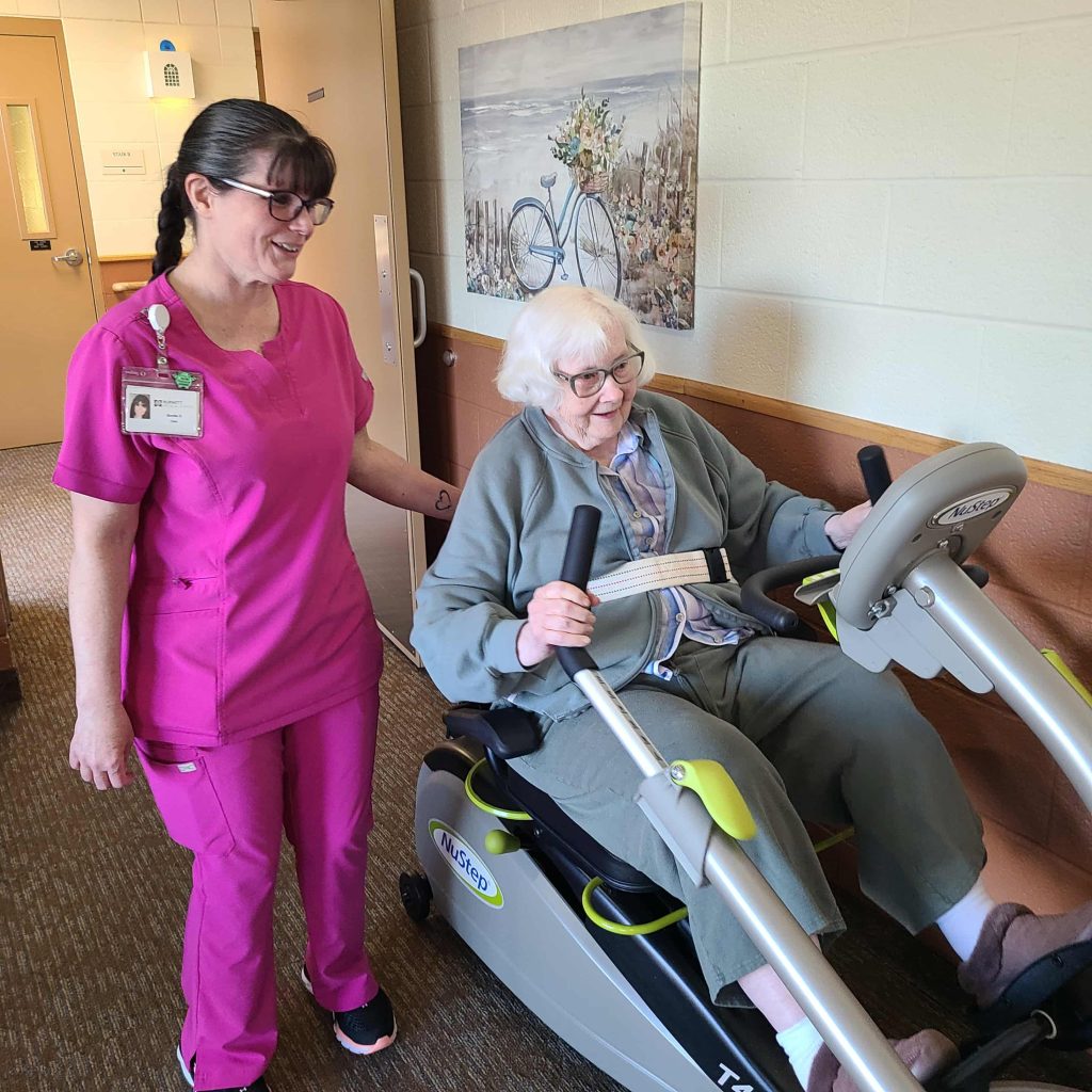 Healthcare professional assisting elderly woman on exercise equipment in Burnett Medical Center's Continuing Care Center, emphasizing patient-centered support and rehabilitation.