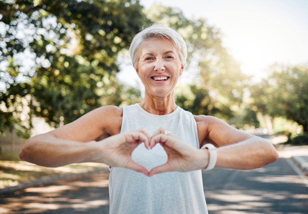 Smiling older woman outdoors forming a heart shape with her hands, promoting wellness and community health, relevant to women's health services at Burnett Medical Center.