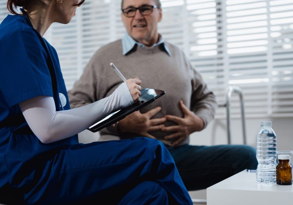 A young Caucasian nurse provides medical consultation to an elderly Caucasian man sitting on a sofa, addressing chronic conditions like obesity, hypertension, diabetes,heart disease, dementia, joint